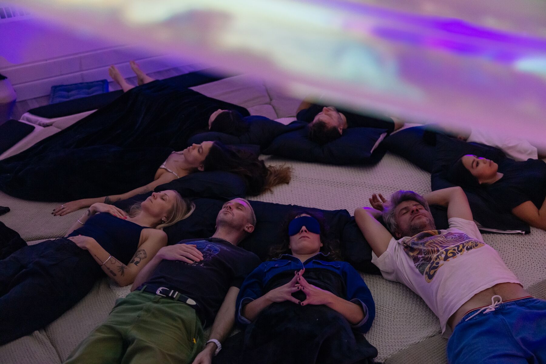 Eight guests lying head to head on AVA, eyes closed, under a sky of projected violet and blue light.
