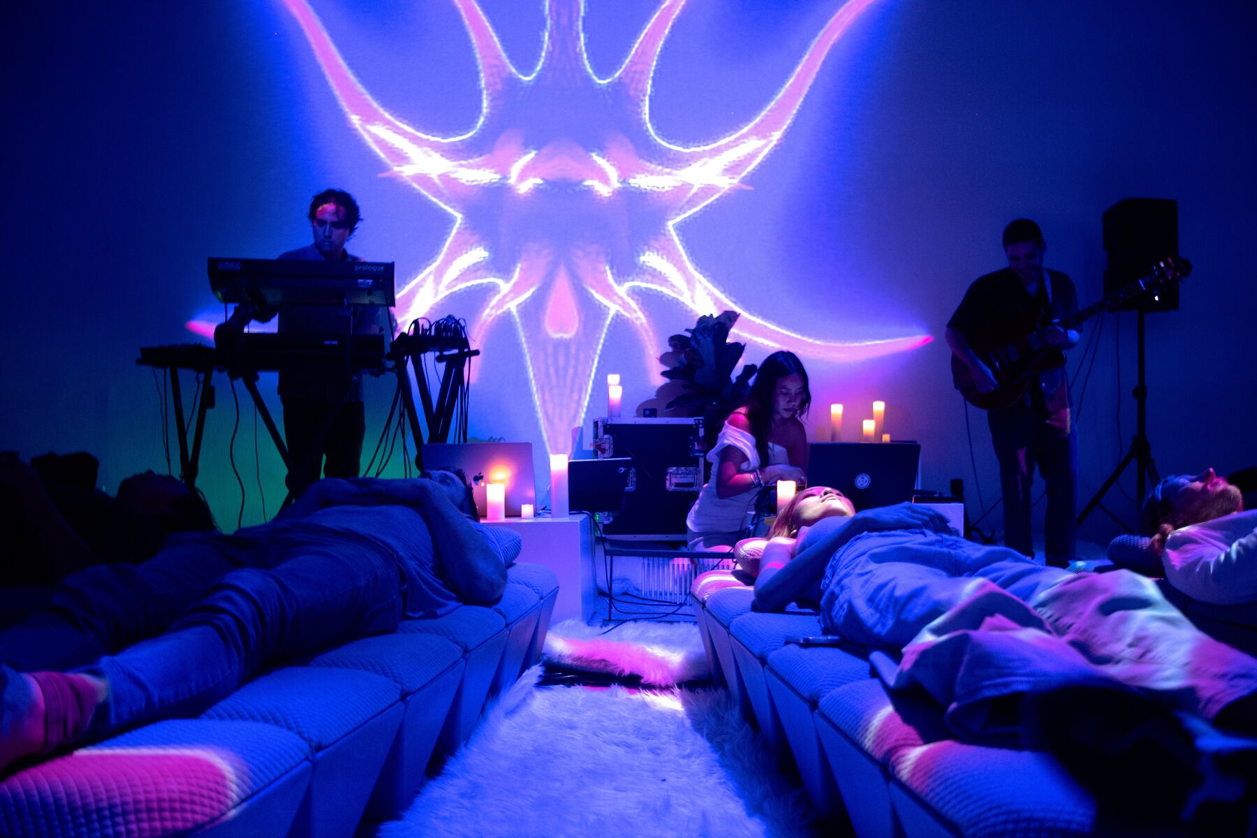 A live set: keys and guitar under a projected visual, guests lying on AVA between candles.